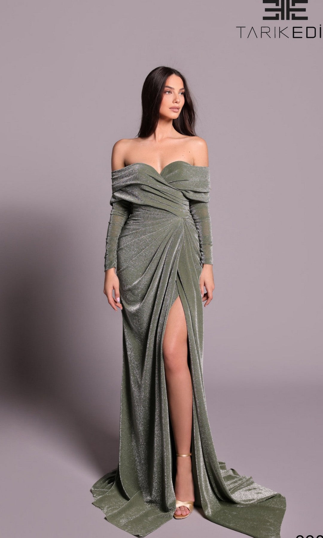 A woman poses against a plain light gray background in the Tarik Ediz 99029 long formal dress, featuring a shimmering nilgreen off-the-shoulder design with a thigh-high slit.